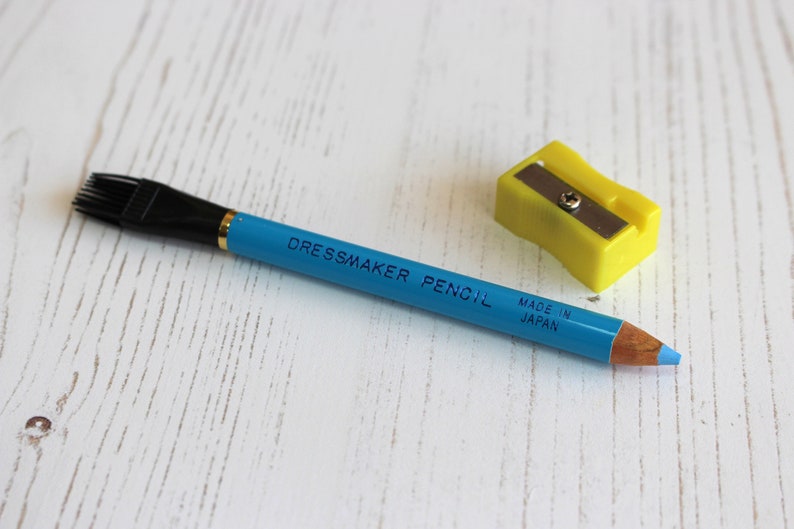 May include: A blue dressmaker pencil with a black brush on one end and the words "Dressmaker Pencil Made in Japan" printed on the side. A yellow pencil sharpener is also pictured.