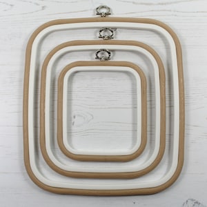 May include: Set of three embroidery hoops with wooden frames and white plastic rings. The hoops are nested inside each other, with the largest hoop at the bottom and the smallest hoop at the top.