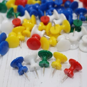 May include: A collection of colorful push pins in red, yellow, blue, green, and white. The push pins have a round head and a pointed end. They are scattered on a white surface.