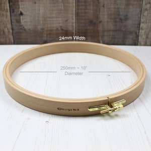 Puede incluir: Un aro de bordado de madera de 250 mm de diámetro y 24 mm de ancho. El aro está hecho de madera natural y tiene un tornillo de apriete de latón. El aro está etiquetado como "Nurge No. 6".