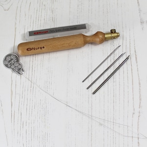 Op de afbeelding: Een houten priknaaldgereedschap met een messing punt en een doorzichtig plastic doosje met het label "Punch Iğnesi / Punch Needle". Het gereedschap wordt geleverd met drie metalen naalden en een metalen draadtrekker met een bloemmotief.