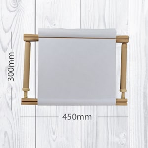 Pode incluir: Um aro de bordado de madeira com uma tela branca esticada sobre ele. O aro tem 450 mm de largura e 300 mm de altura.