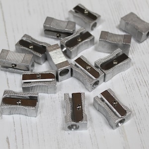 May include: A group of 12 silver pencil sharpeners on a white wooden surface. The sharpeners are all the same design, with a rectangular body and a round hole for the pencil. They have a silver finish and a black plastic blade.
