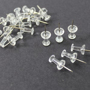 May include: A collection of clear, plastic thumb tacks with silver metal pins. The tacks are scattered on a dark gray surface, with some grouped together and others spread out.