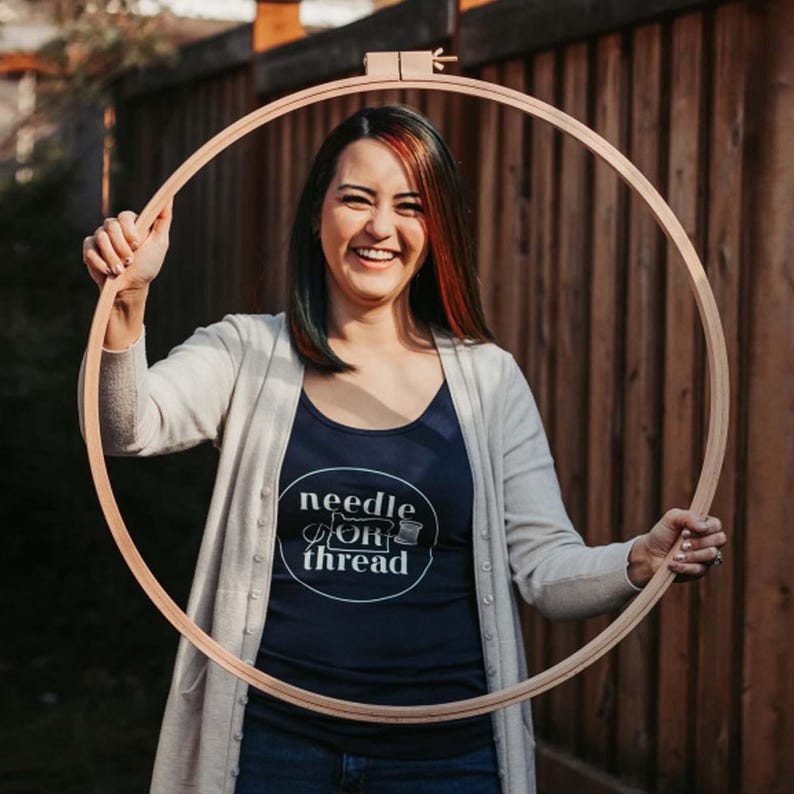May include: A woman wearing a navy blue tank top with the text "needle & thread" in a white circle. She is holding a large wooden embroidery hoop in front of her.