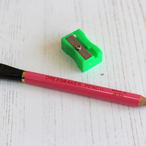 May include: A pink dressmaker pencil with a brush on one end and a sharpener on the other. The pencil is labeled "Dressmaker Pencil" and "Made in Japan".