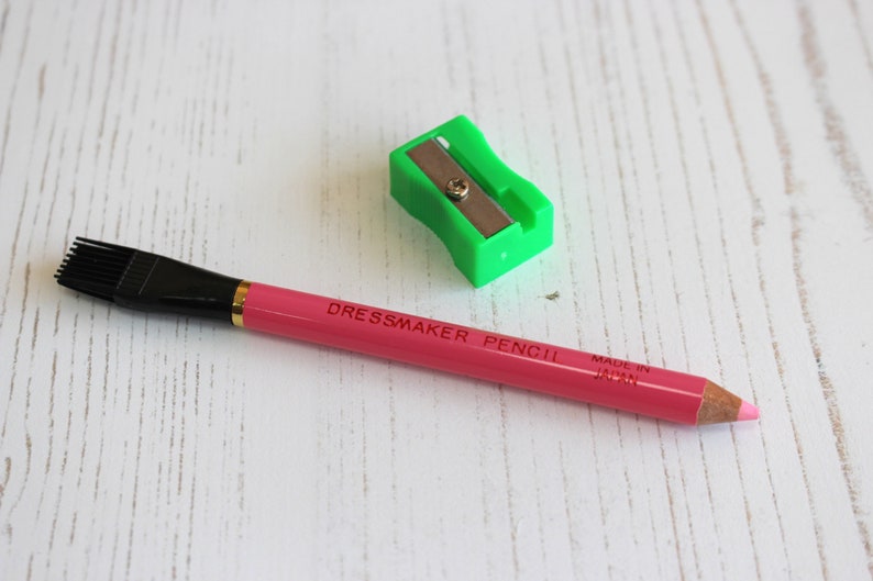 May include: A pink dressmaker pencil with a brush on one end and a sharpener on the other. The pencil is labeled "Dressmaker Pencil" and "Made in Japan".