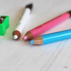 May include: Three sharpened wooden pencils with different colored leads: white, pink, and blue. The pencils are lying on a white surface.