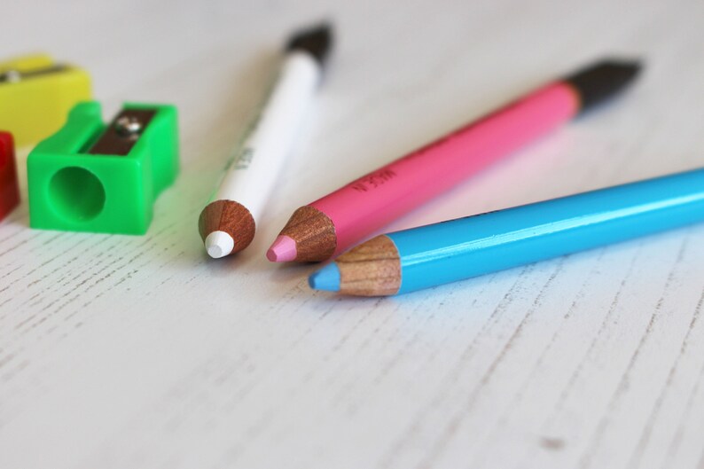 May include: Three sharpened wooden pencils with different colored leads: white, pink, and blue. The pencils are lying on a white surface.