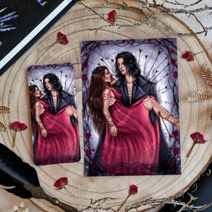 Könnte beinhalten: Zwei Kunstdrucke mit einem Paar in einer romantischen Umarmung, umgeben von Pfeilen und Rosen. Die Frau trägt ein rotes Kleid, und der Mann ist in einem schwarzen Mantel. Die Drucke liegen auf einer Holzoberfläche mit getrockneten Blumen und einem Buch.