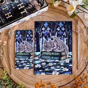 May include: A rectangular bookmark and a matching rectangular art print featuring a sleeping gargoyle resting on two stacked books titled The Knave and the Moon and The Knight and the Moth by Rachel Gillig, surrounded by white flowers in dark water.