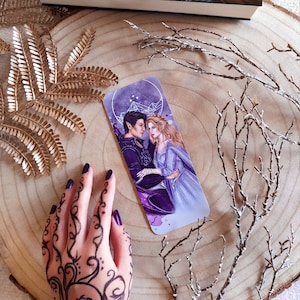 May include: A purple and blue bookmark featuring a couple, a man with dark hair and a woman with long blonde hair, standing in front of a starry sky with a geometric design.