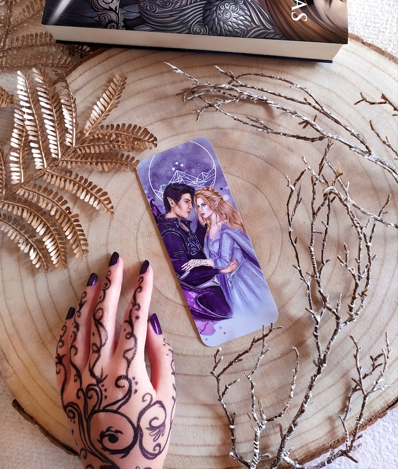 May include: A purple and blue bookmark featuring a couple, a man with dark hair and a woman with long blonde hair, standing in front of a starry sky with a geometric design.