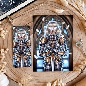 Op de afbeelding: Illustratie van een vrouw in zilveren en gouden harnas, met een zwaard, met een vlindermasker. Het kunstwerk staat op een bladwijzer en een grotere print. De achtergrond toont een glas-in-loodraam en vlinders. Een boek getiteld "Night Moth" is zichtbaar.