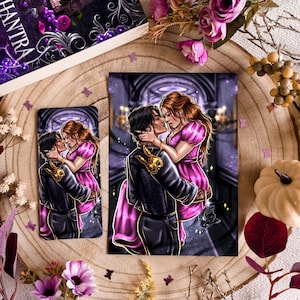 Puede incluir: Una impresión artística y una funda de teléfono con una ilustración romántica de una pareja. La mujer lleva un vestido rosa, y el hombre una chaqueta negra con detalles dorados. La portada del libro muestra el título "Enchantra" con una calavera y motivos florales.