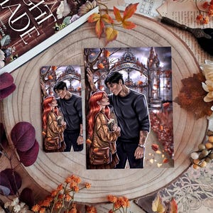 May include: Two art prints and a book are displayed on a wooden surface. The prints depict a couple in an autumn setting. The book cover reads "Night Shade". The scene is decorated with fall foliage and flowers.