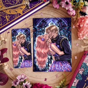 May include: An illustration of a couple kissing, with the woman having long pink hair and wearing a purple dress. The man is wearing a black jacket. The image is on a postcard and a smaller rectangular item, surrounded by flowers and a book.