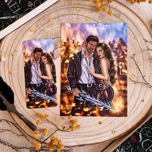 May include: A print and bookmark featuring a couple embracing, set against a fiery background. The man holds a glowing sword. The artwork is on a wooden surface with floral accents and a book titled "The Book of Azrael".