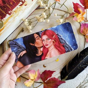 Puede incluir: Un marcap&aacute;ginas con una pareja con alas, la mujer tiene el pelo rojo y lleva un collar de oro, el hombre tiene el pelo oscuro y lleva una camisa negra. El fondo es un cielo azul con rayos.