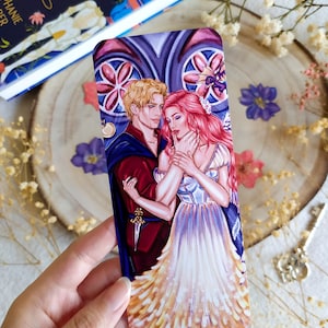 May include: A bookmark featuring a romantic illustration of a couple in a gothic setting. The man is wearing a red and blue outfit, while the woman is wearing a white dress with a pink bodice. The background is a stained glass window with a floral design.