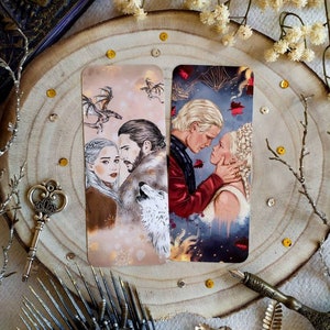 Op de afbeelding: Twee bookmark-ontwerpen met een stel uit een populaire fantasy-televisieserie. De eerste bookmark toont een stel dat voor een witte wolf staat met een draak die erboven vliegt. De tweede bookmark toont een stel dat voor een vurige achtergrond staat met vallende rode bladeren.