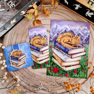 Puede incluir: Un dragón dorado enroscado en una pila de libros con los títulos "A Court of Thorns and Roses", "A Court of Mist and Fury", "A Court of Wings and Ruin", "A Cruel Prince" y "The Fourth Wing". Los libros están sobre una superficie verde cubierta de hierba con rosas rojas.