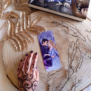 May include: A bookmark featuring a fantasy novel cover with two characters, a man and a woman, standing together. The bookmark is on a wooden surface with gold and white branches.