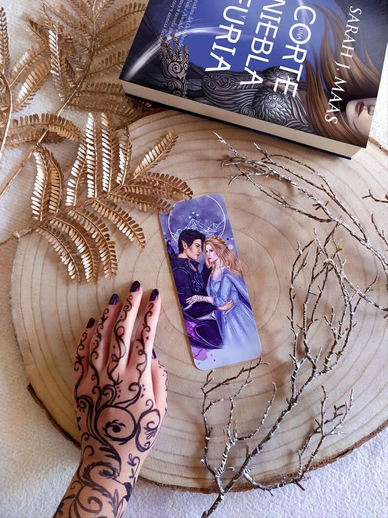 May include: A bookmark featuring a fantasy novel cover with two characters, a man and a woman, standing together. The bookmark is on a wooden surface with gold and white branches.