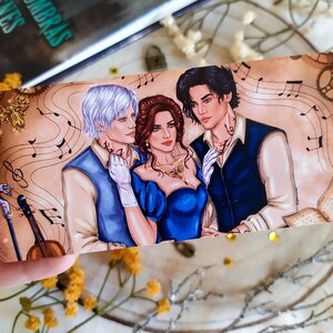 May include: A bookmark featuring a romantic illustration of three people, two men and one woman, with musical notes and a vintage aesthetic. The woman is wearing a blue dress and the men are wearing blue and white shirts. The bookmark has a gold background with a gold gear design.