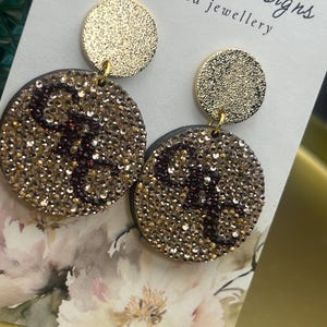 May include: Gold-toned, handcrafted earrings with a circular design. The earrings feature a smaller, solid gold circle at the top and a larger, embellished circle below, covered in gold and dark brown rhinestones. The text "Mia May Designs handcrafted jewellery" is visible.