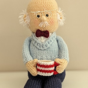 Può includere: Una bambola di lana con un maglione blu, un papillon rosso e occhiali, che tiene una tazza a righe rosse e bianche.