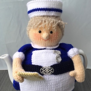 Puede incluir: Una funda de tetera de punto con forma de enfermera. La funda es blanca con detalles azules y un cinturón negro. La enfermera lleva una gorra blanca con una banda azul.