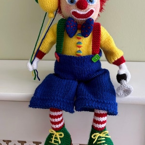 May include: A knitted clown doll with red hair, a red nose, and a painted smile. The clown wears a yellow top, blue shorts, and green shoes. It holds yellow balloons and a small grey object. The doll has a colourful bow tie and braces.