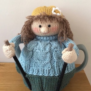 Puede incluir: Una funda de tetera de punto en forma de persona que lleva un suéter azul, pantalones verdes, un sombrero amarillo y una flor blanca. La persona sostiene dos agujas de tejer.