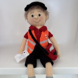 May include: Hand-knitted doll of a postman, wearing a black cap, red shirt, orange vest with reflective stripes, and black shorts. The doll has a satchel and holds a small package with text. Gray and black shoes complete the outfit.