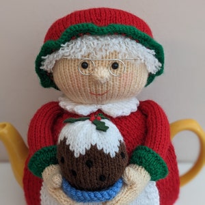 May include: Knitted teapot cozy featuring a smiling woman wearing a red dress, white hat, and glasses, holding a brown and white Christmas pudding.