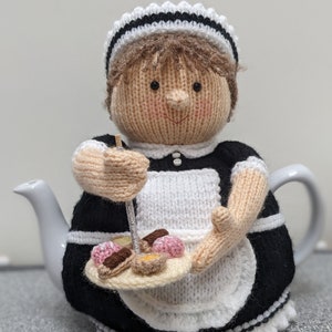May include: A knitted teapot cozy in the shape of a maid holding a tray of pastries. The cozy is black and white with a white apron and a black and white striped hat.