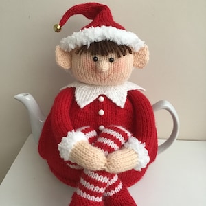 Puede incluir: Una funda de té de punto en forma de elfo de color rojo y blanco con ribete de piel blanca. El elfo está sentado en una tetera con las piernas cruzadas. El elfo lleva un gorro rojo con ribete de piel blanca y una campana dorada.