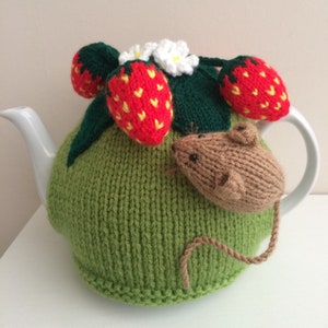May include: A green knitted teapot cosy with a brown knitted mouse and red strawberries with yellow seeds. The cosy is on a white teapot.