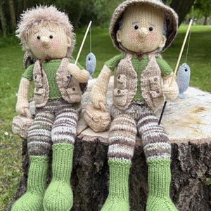 May include: Two knitted dolls dressed in fishing vests and trousers, wearing green socks and hats, holding fishing rods with small blue fish attached. The dolls are sitting on a tree stump.