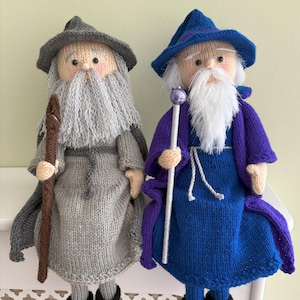 May include: Two knitted wizard dolls. One wears a grey hat, robe, and cape, holding a brown staff. The other wears a blue hat, robe, and purple cape, holding a silver staff with a purple orb. Both dolls have white beards and black shoes.