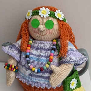 May include: A knitted tea cozy in the shape of a hippie girl with long orange hair, wearing a purple and white striped sweater, a beaded necklace, a green headband with white flowers, and green sunglasses. The cozy has a brown skirt and a green bag with a white flower.