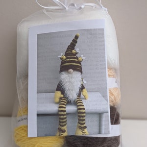 May include: A clear bag tied with a white ribbon contains a gnome craft kit. The kit includes yarn in yellow, brown, and cream colours, and a card with a picture of a gnome wearing a brown and yellow striped hat with bee accents.