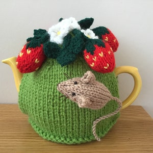 May include: A green knitted teapot cosy with a mouse and strawberries. The cosy has a white flower and green leaves on top. The teapot is yellow.