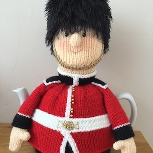 Puede incluir: Una funda de tetera de punto con forma de guardia británico. La funda es roja con ribetes negros y botones dorados. El guardia lleva un gorro de piel de oso negro y tiene la cara blanca.