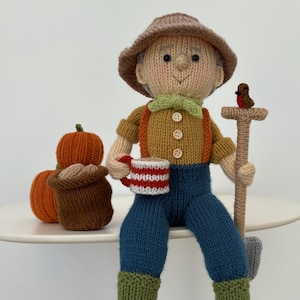 Puede incluir: Figura de granjero tejida a mano, con sombrero marrón, camisa color mostaza, pantalones azules y botas verdes. El granjero sostiene una taza a rayas rojas y blancas y una pequeña pala. Hay calabazas tejidas cerca.