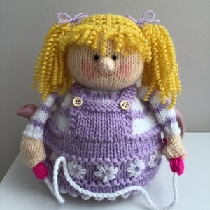 Puede incluir: Una funda de tetera de punto en forma de niña con cabello amarillo, vestido morado y detalles blancos. La funda tiene una cuerda rosa atada.