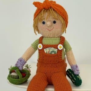 Puede incluir: Muñeca tejida a mano que representa a un jardinero, con un mono naranja, una camisa verde y un pañuelo naranja. La muñeca sostiene una cesta verde con verduras y un pepino verde. La muñeca tiene pelo rubio y ojos azules. El mono tiene botones en forma de margarita.