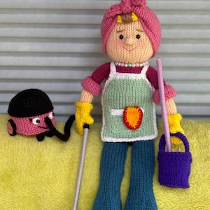 Op de afbeelding: Handgemaakte gebreide pop die een schoonmaakster voorstelt, met een roze hoofddoek, een groene schort en een blauwe broek. Ze houdt een dweil en een paarse emmer vast. Een roze stofzuiger staat naast haar. De pop staat op een gele ondergrond.