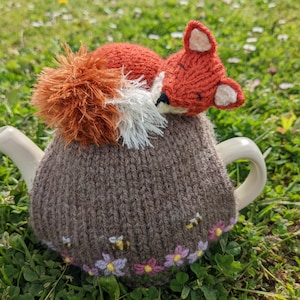 Könnte beinhalten: Ein brauner gestrickter Teekannenwärmer mit einem Fuchsdesign. Der Fuchs ist orange und weiß mit einem flauschigen Schwanz. Der Wärmer ist mit gestickten Blumen und Bienen am unteren Rand verziert.